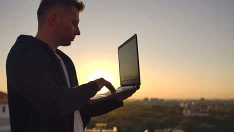 Hacker-using-laptop-on-rooftop-with-city-view-and-forex-chart.-Hacking-and-stats-concept.-A-man-at-sunset-in-slow-motion-writing-software-code-on-a-laptop